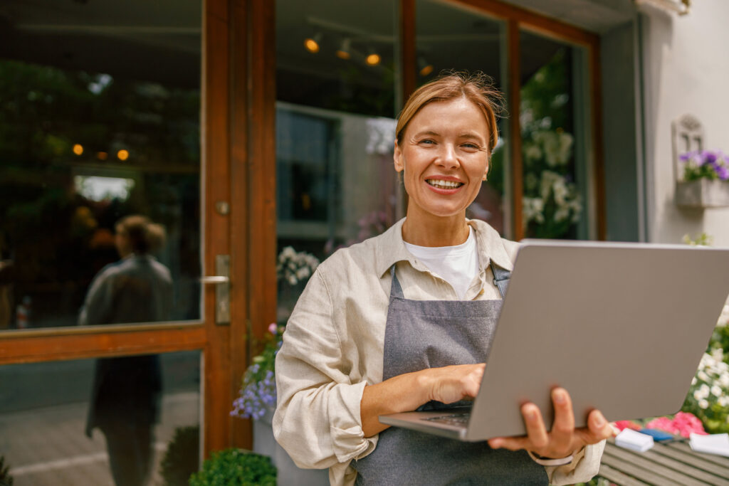Smiling flower shop owner holding a laptop outside her storefront, representing a small business managing its Google Business Profile category